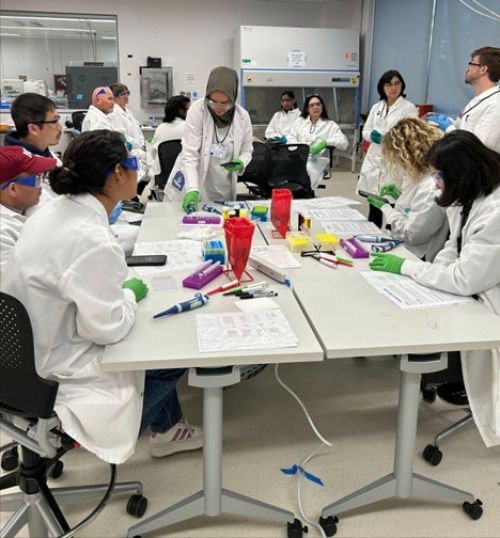 Numerous people in white lab coats standing around white tables working on biotech lab projects
