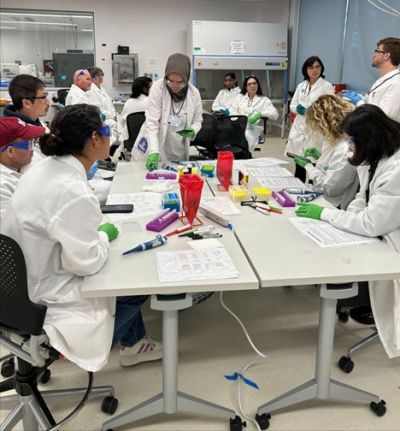 Numerous people in white lab coats standing around white tables working on biotech lab projects